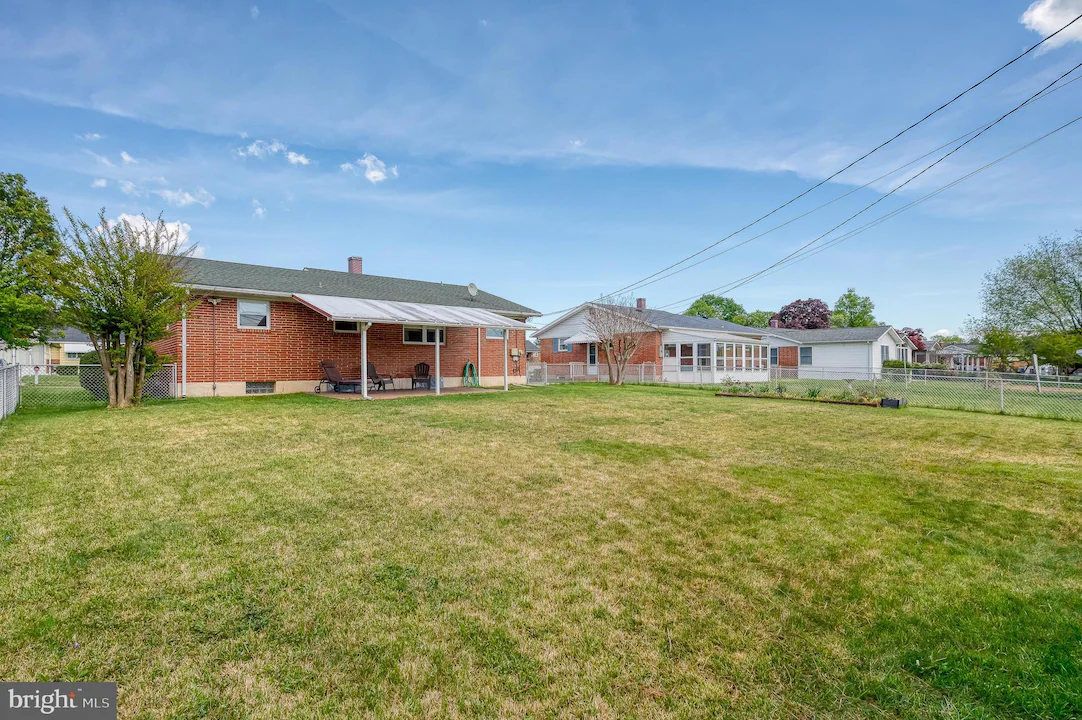 Backyard of a single-story brick house with a covered patio, outdoor seating, and a lawn bordered by a chain-link fence under a blue sky
