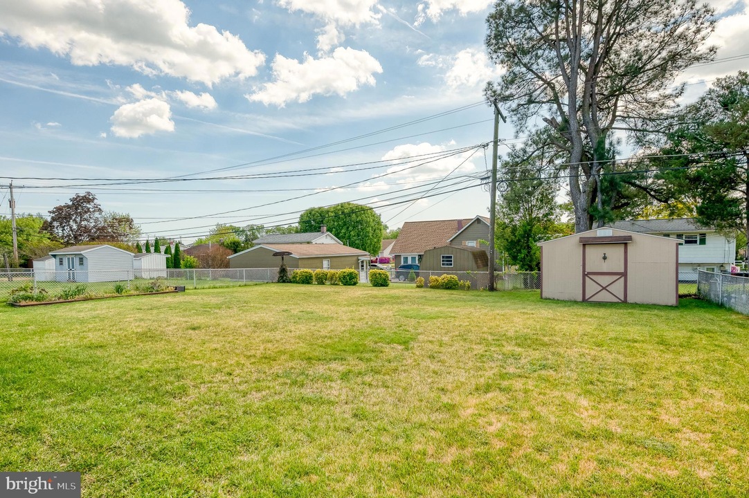 Suburban backyard with a beige shed, chain-link fence, and trimmed lawn, facing neighboring houses and utility poles under a blue, partly cloudy sky