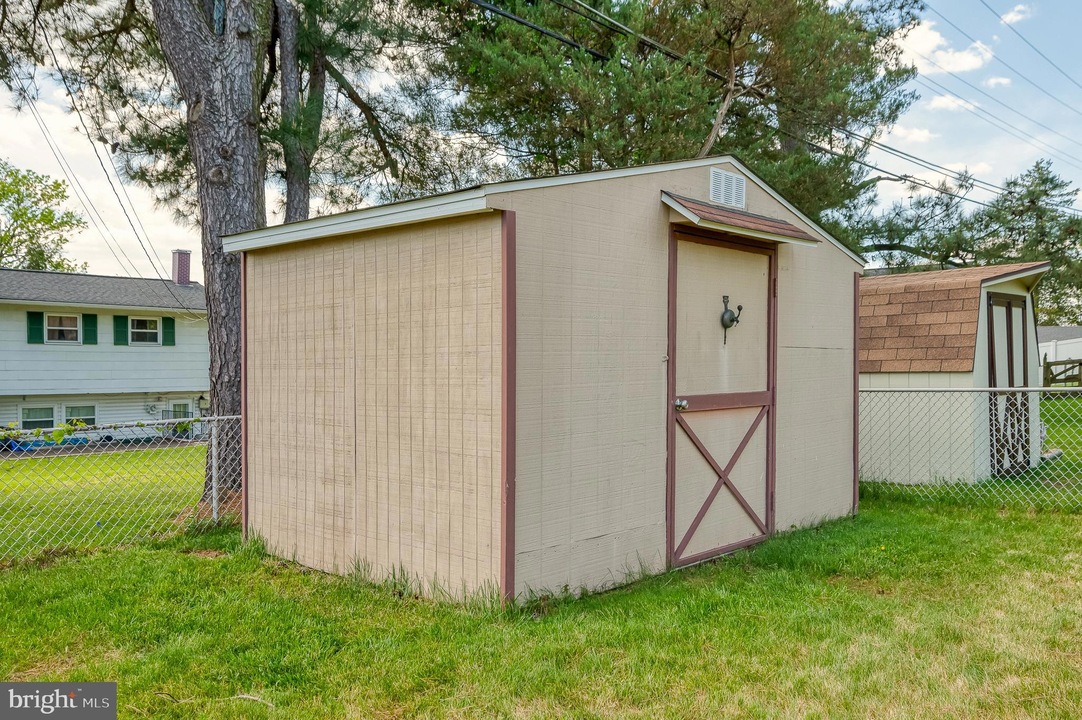 Beige storage shed with brown trim and a cross-braced door in a fenced residential backyard, with a large tree and neighboring houses in the background.