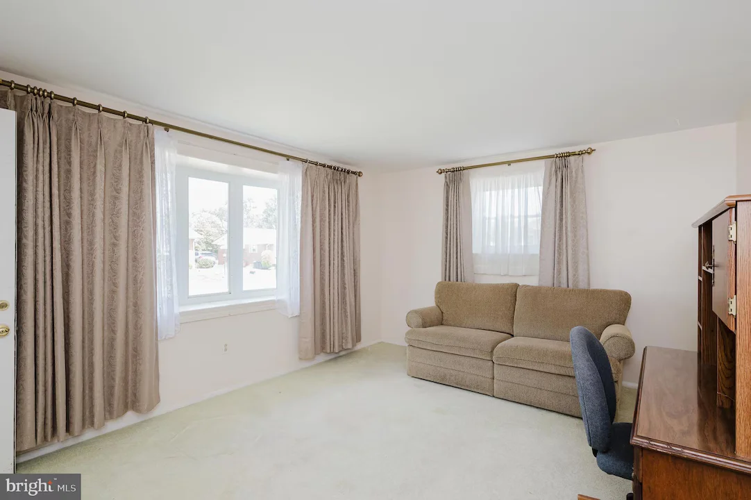 Living room featuring a beige sofa, a wooden desk with a chair, and large windows with heavy drapes.