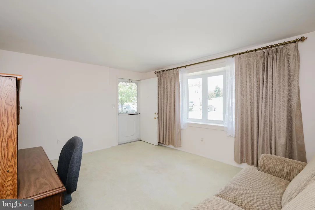 Living room with a beige sofa, wooden desk and chair, white walls, and large window with sheer and beige curtains.