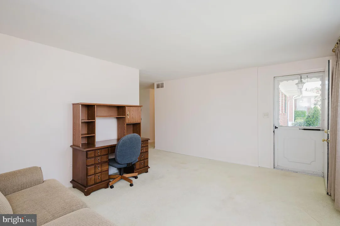 Wooden desk with built-in shelves and a blue rolling office chair in a sparsely furnished living room with light walls and a door outside.