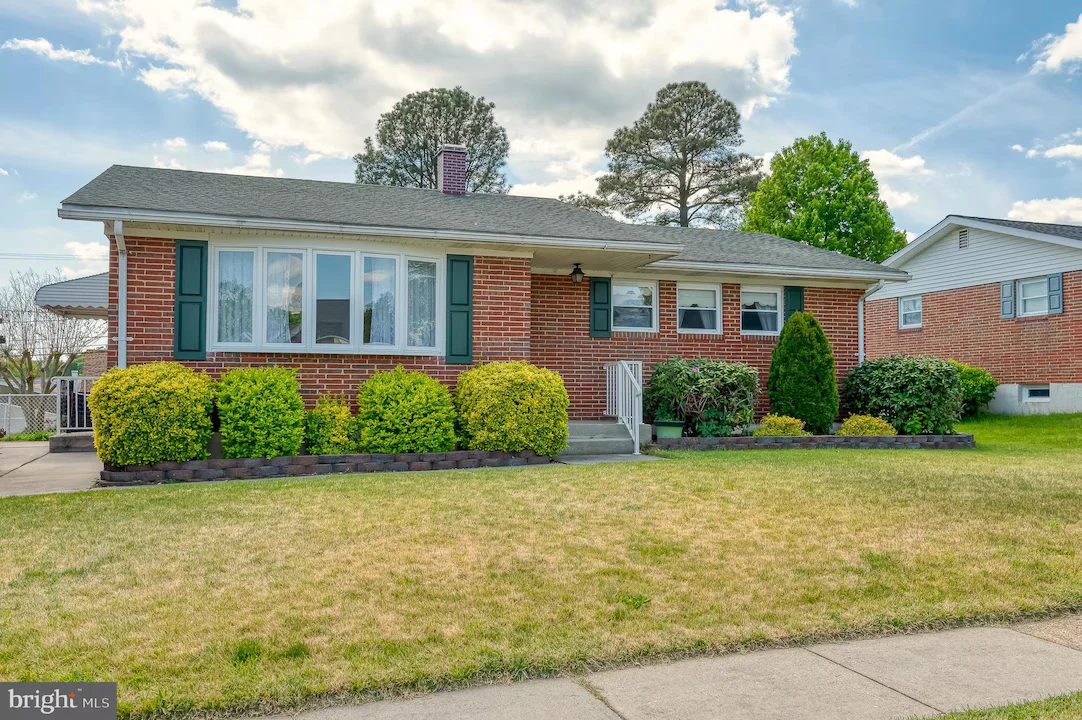 Single-story brick house with green shutters and a manicured front garden of shrubs and small trees.