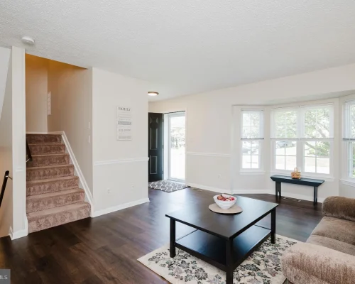 Bright living room with dark wood floors, beige sofa, black coffee table on a patterned rug, and a bay window; stairs lead to the left behind a wall.
