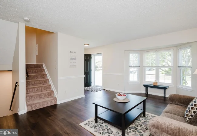 Bright living room with dark wood floors, beige sofa, black coffee table on a patterned rug, and a bay window; stairs lead to the left behind a wall.
