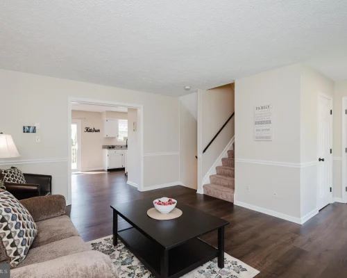Living room with a brown sofa, patterned throw pillows, a black coffee table, and a lamp; doorway opens to a white kitchen.