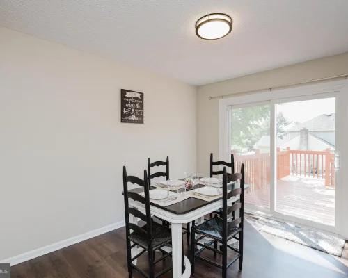 Dining area with a square table set for four and black chairs, next to a sliding glass door opening to a wooden deck outside.