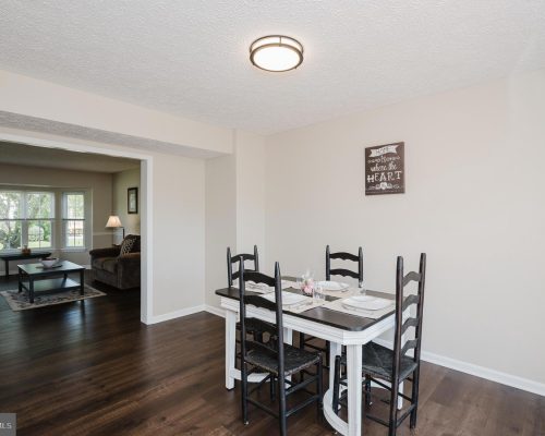 Dining area with a white table set for four and dark chairs, beige walls, and a wall sign reading 'Home is where the heart is' with a living room visible beyond the doorway.