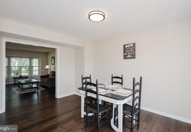 Dining area with a white table set for four and dark chairs, beige walls, and a wall sign reading 'Home is where the heart is' with a living room visible beyond the doorway.