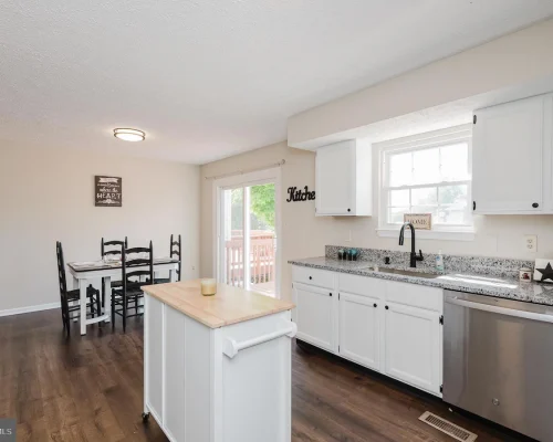 Bright kitchen with white cabinets, granite countertops, island, and a stainless dishwasher, adjacent to a dining table area.
