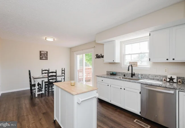 Bright kitchen with white cabinets, granite countertops, island, and a stainless dishwasher, adjacent to a dining table area.