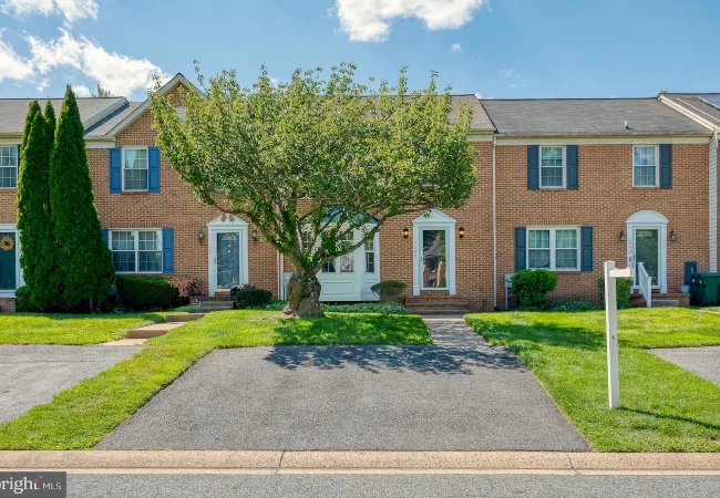 Brick townhouses with blue shutters line a sunny suburban street; a large tree dominates the front yard of the center home, with driveways and well-kept lawns nearby.