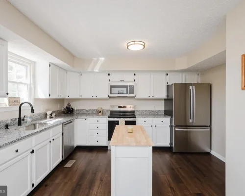 Bright kitchen with white cabinets, granite countertops, and a central island; stainless steel fridge and stainless appliances along the back wall.