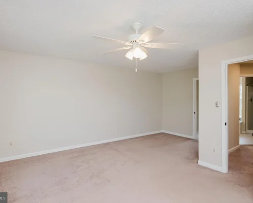 Empty beige bedroom with a white ceiling fan and light, beige carpet, and a doorway to an adjacent bathroom on the right.