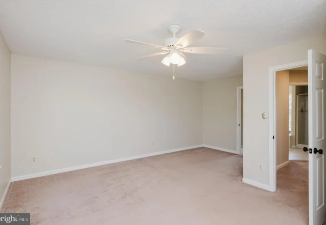 Empty beige bedroom with a white ceiling fan and light, beige carpet, and a doorway to an adjacent bathroom on the right.