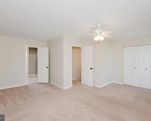 Empty bedroom with beige walls, light carpet, and a white ceiling fan; open doors reveal a bathroom and a hallway, with a double-door closet on the right.