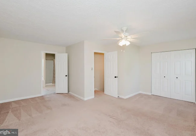 Empty bedroom with beige walls, light carpet, and a white ceiling fan; open doors reveal a bathroom and a hallway, with a double-door closet on the right.