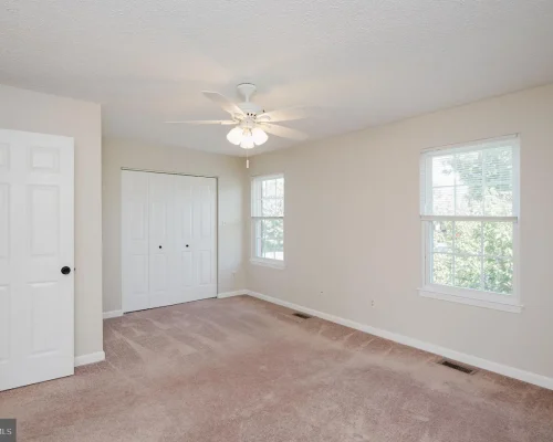 Vacant bedroom with beige walls, pinkish carpet, a white ceiling fan, a double-door closet, and two windows letting in natural light.