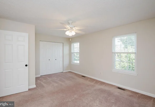 Vacant bedroom with beige walls, pinkish carpet, a white ceiling fan, a double-door closet, and two windows letting in natural light.