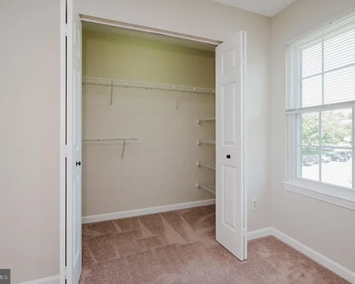 Empty beige closet with white wire shelves and carpeted floor, open double doors beside a window.