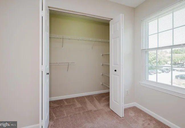 Empty beige closet with white wire shelves and carpeted floor, open double doors beside a window.