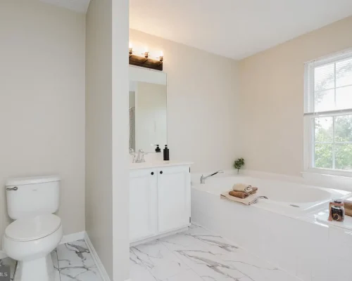 Bright modern bathroom with a white vanity, rectangular mirror, and a large soaking tub by a window; tiled marble floor throughout.