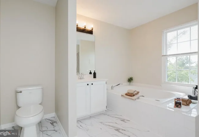 Bright modern bathroom with a white vanity, rectangular mirror, and a large soaking tub by a window; tiled marble floor throughout.
