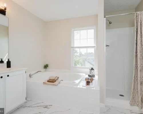 Bright bathroom with a white corner vanity, large bathtub, and a separate shower with a patterned curtain; window lets in natural light.
