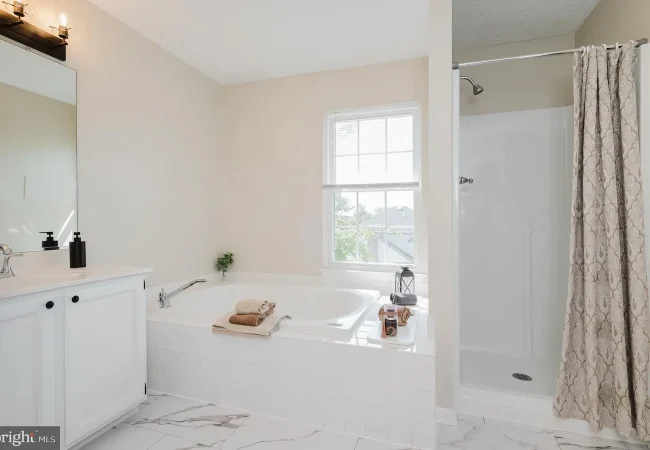 Bright bathroom with a white corner vanity, large bathtub, and a separate shower with a patterned curtain; window lets in natural light.
