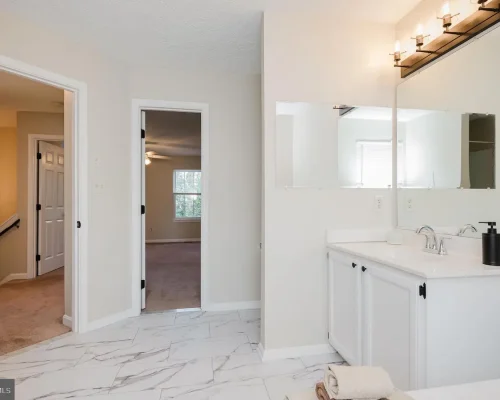 Bright bathroom with white cabinetry and a double sink, large mirrored vanity, and a four-bulb light fixture. Marble floor and beige walls.
