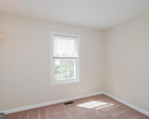 Empty beige room with a white-framed window and blinds; a floor lamp in the left corner.
