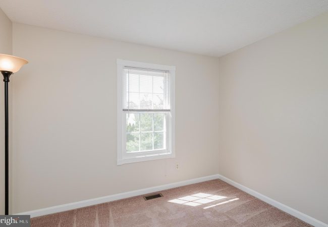 Empty beige room with a white-framed window and blinds; a floor lamp in the left corner.