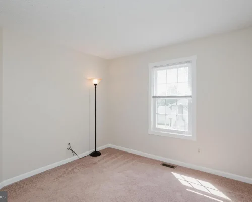 Empty beige-carpeted bedroom with a single floor lamp, a window with blinds, and white baseboard trim.