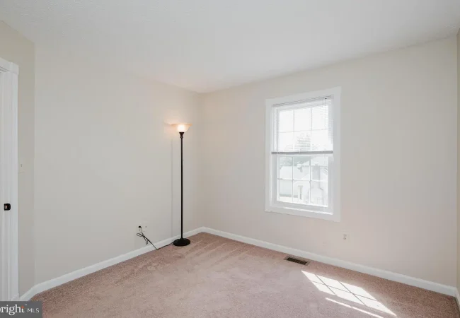 Empty beige-carpeted bedroom with a single floor lamp, a window with blinds, and white baseboard trim.