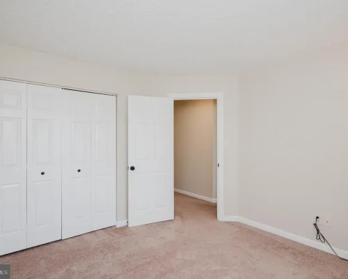 Empty beige bedroom with light brown carpet, a double closet with white doors on the left, and an open doorway to a hallway on the right; a wall sconce provides light.
