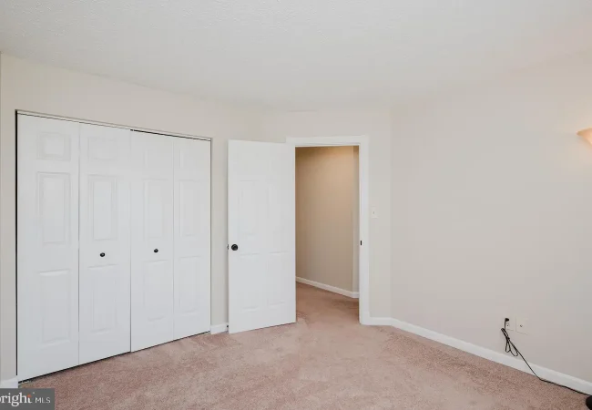 Empty beige bedroom with light brown carpet, a double closet with white doors on the left, and an open doorway to a hallway on the right; a wall sconce provides light.