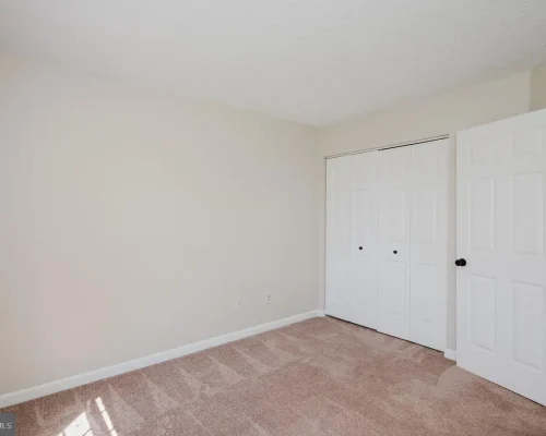 Empty beige bedroom with light carpet, white closet doors, and an open entry door to another room.