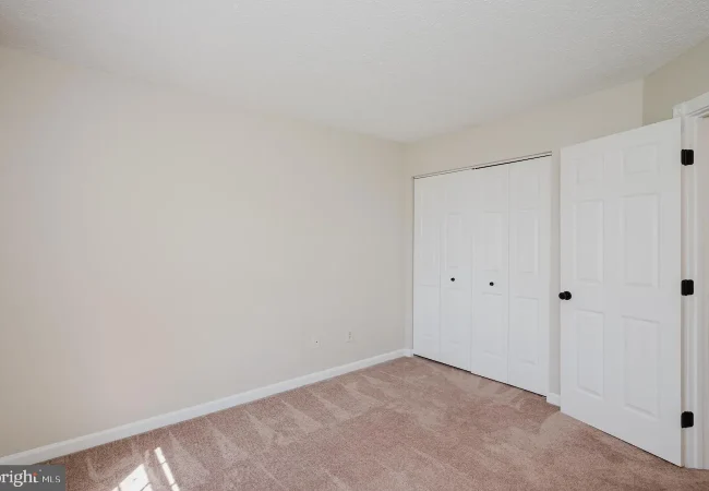 Empty beige bedroom with light carpet, white closet doors, and an open entry door to another room.