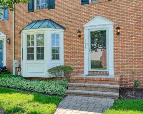 Brick townhouse facade with three entrances: a blue door on the left, a white bay-windowed nook in the center, and a glass door on the right, with a manicured lawn and shrubs nearby.