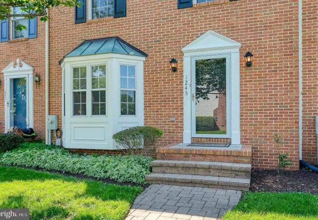 Brick townhouse facade with three entrances: a blue door on the left, a white bay-windowed nook in the center, and a glass door on the right, with a manicured lawn and shrubs nearby.