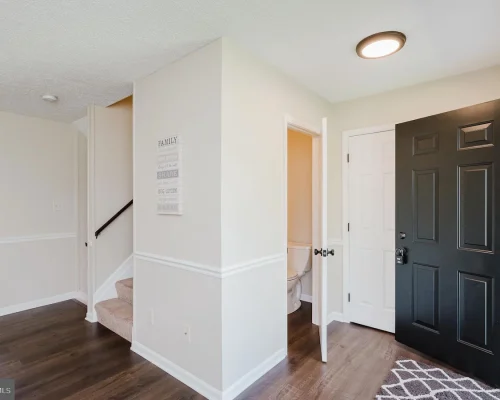 Entryway with a black front door open to a hallway; white walls, wood floor, and stairs to the left.