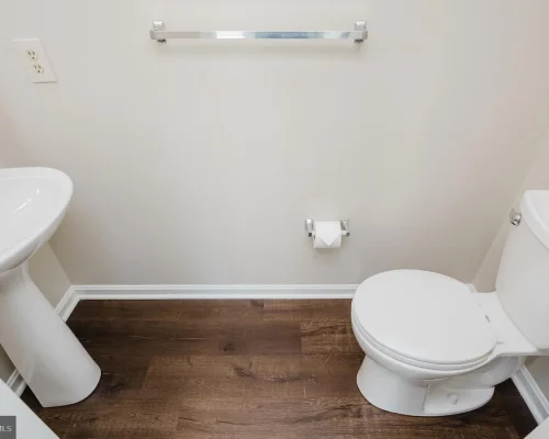 Small bathroom with a white pedestal sink on the left, white toilet on the right, chrome towel bar, toilet paper holder, and dark wood-look flooring.