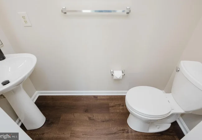 Small bathroom with a white pedestal sink on the left, white toilet on the right, chrome towel bar, toilet paper holder, and dark wood-look flooring.