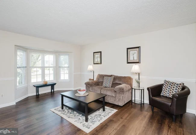 Living room with beige sofa, brown leather chair, coffee table, and bay window area in a bright, neutral decor.