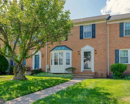 Brick townhouse with blue shutters and a white bay window, set in a green front yard with a large tree and brick walkway leading to the front door.