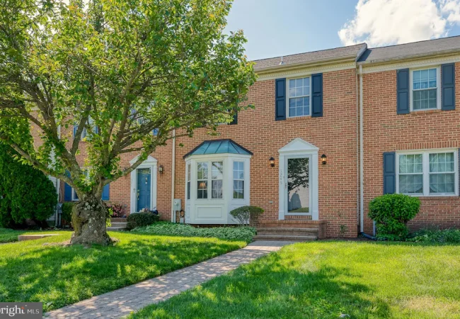 Brick townhouse with blue shutters and a white bay window, set in a green front yard with a large tree and brick walkway leading to the front door.