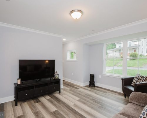 Living room with a flat-screen TV on a black media console, beige sofa, and a large window letting in daylight outside edge light fixture above.