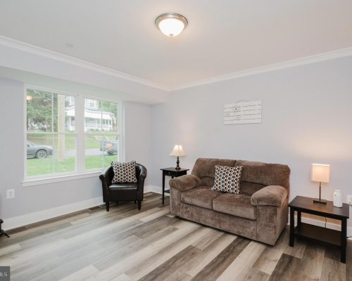 Bright living room with light gray walls, a brown sofa with patterned cushions, a black armchair by a window, two dark wood side tables with lamps, and a small wall art piece; light wood flooring