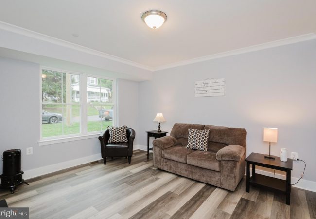 Bright living room with light gray walls, a brown sofa with patterned cushions, a black armchair by a window, two dark wood side tables with lamps, and a small wall art piece; light wood flooring