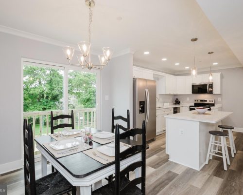 Bright open dining and kitchen area with a white table set for four, black chairs, and a chandelier near a sliding glass door to a green backyard.